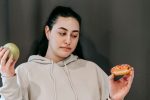 Contemplative plump female in hoodie choosing between ripe green apple and delicious donut in studio