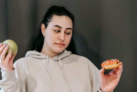 Contemplative plump female in hoodie choosing between ripe green apple and delicious donut in studio