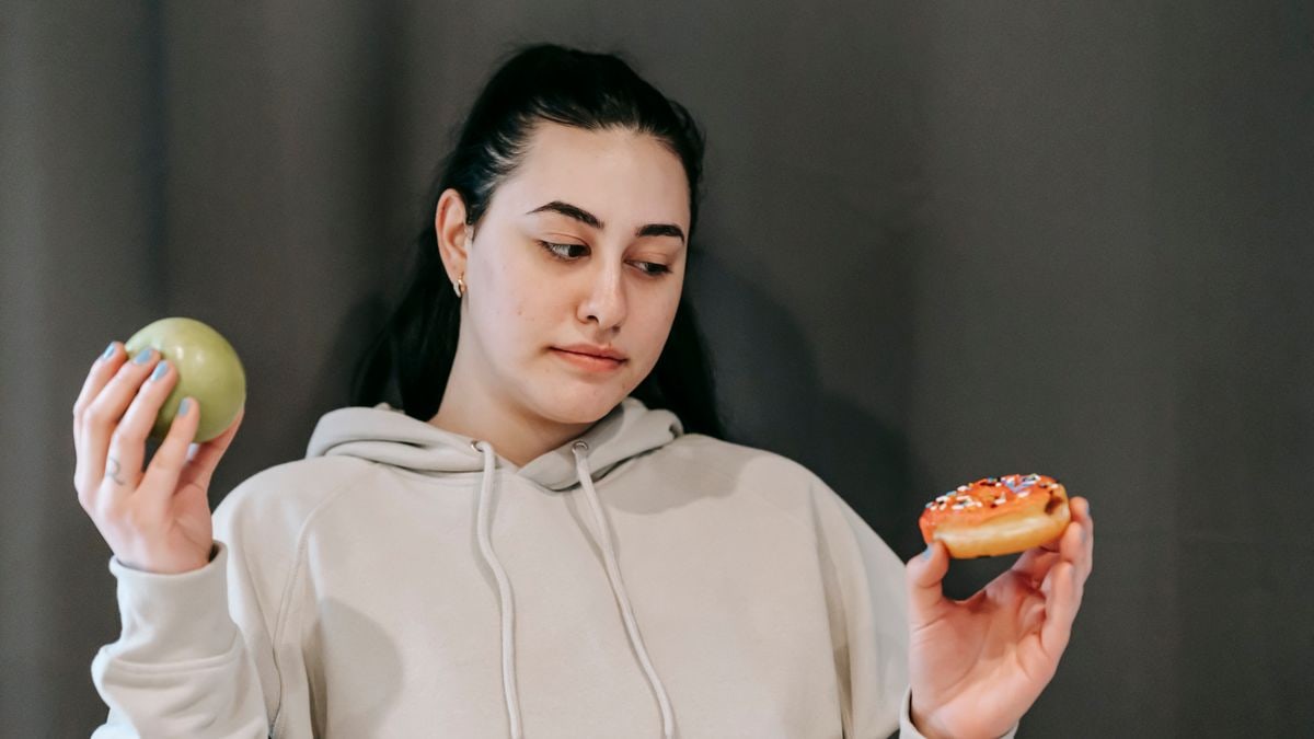 Contemplative plump female in hoodie choosing between ripe green apple and delicious donut in studio